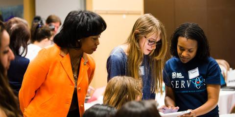 Photo of three women at a conference, observing something on a tablet.