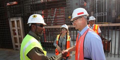 Two workers in hard hats and safety vets shake hands on a construction site.